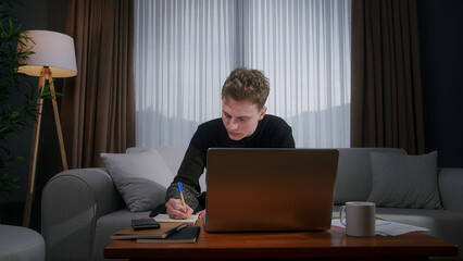 Young blonde man working from home using with laptop while sitting on sofa at home. Focused millennial male looking at laptop, taking note on notebook	
