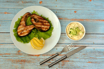 Two grilled salmon steaks with lettuce, lemon and sauce on a wooden table.