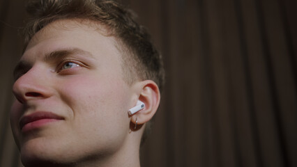 Close-up of young blonde man with earrings wearing wireless in-ear headphones and listening to music at home	
