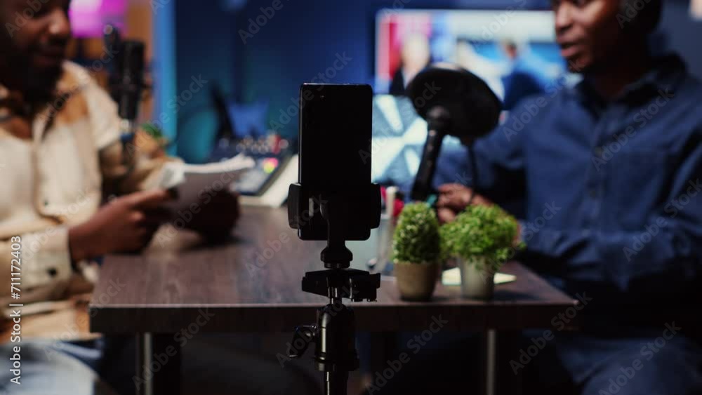 Poster focus on smartphone on tripod used by podcast host in blurry background to record guest telling inte