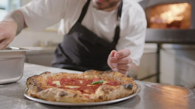 Close-up view of male chef adding leaves of fresh basil to just baked delicious pizza with salami and bubbly crust before serving it to customers in pizzeria