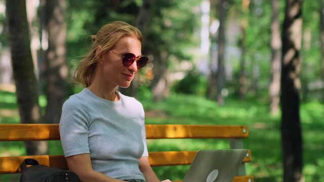 Relaxed blonde woman sitting on the bench using a laptop, close up. Cute female freelancer making remote work in the summer city park on blurred green foliage background.