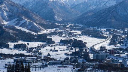 Panoramic view of a quaint town nestled in a valley with snow-covered fields and surrounding mountain peaks.