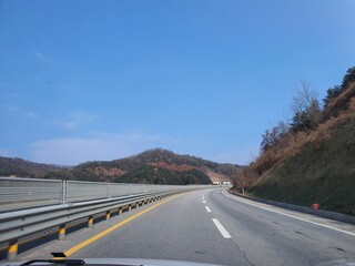 A straight road stretches into the distance under a clear blue sky. Mountains rise on either side, framing the road. The scene is bright and open, with the road leading the eye towards the horizon.