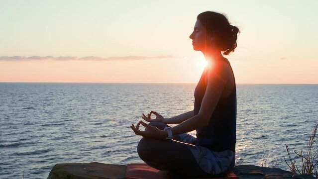 Woman meditating with closed eyes sits in lotus pose Prithvi Mudra sea sunset. Female sitting on shore practicing yoga admiring nature. Sport recreation, mental spirit, practice, inner life concept.
