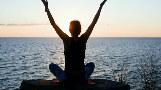 Silhouette woman meditating folding hands in namaste sitting in lotus pose by sea at sunset. Female on shore practicing yoga admiring wild nature. Rear view. Mental spirit, practice concept.