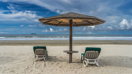 Two sun loungers with soft mattresses stand on the sandy beach under a sun umbrella. Ahead is the turquoise ocean. The waves are rolling towards the shore. There's a ship on the horizon. Malaysia. 
