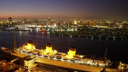 Aerial Panning Shot Of The Queen Mary Cruise Ship Moored In Sea By Illuminated City Against Sky At Night - Long Beach, California
