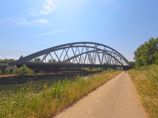 This image features a modern steel arch bridge spanning a calm river, captured on a bright summer day. The perspective is taken from a paved path that parallels the river, inviting the viewer into the