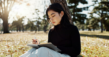 Japanese woman, writing and book in park, thinking and relax by tree, grass or sunshine for peace. Girl, person or student with story in nature, college or notes with vision, knowledge or campus lawn
