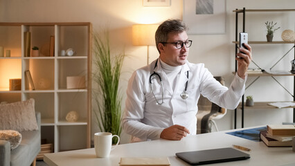 Fototapeta na wymiar Medical video advice. Health consultation. Online meeting. Healthcare professional man talking at virtual conference on smartphone sitting at desk in clinic interior.