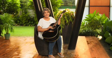 Asian Indian Hindu happy gen x older middle aged woman lady sitting chair singing song play guitar...