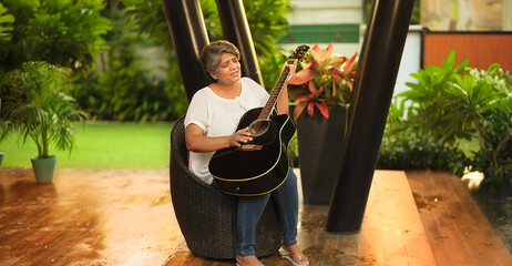 Asian Indian Hindu happy gen x older middle aged woman lady sitting chair singing song play guitar...