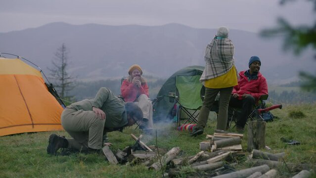 Multiethnic group of tourists rests in camp in forest. Caucasian man blows campfire. Adult woman sits, talks with hiking buddies and takes photos on phone. Hikers during their vacation to mountains.
