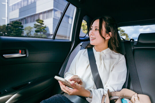 スマホを持ちながら自動車の後部座席で窓の外を眺めるビジネスウーマン（出張・検索・アプリ）
