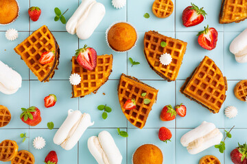 Sweet waffles, meringues, cupcakes and fresh strawberries on a blue tile background. Top view.