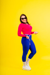 Pretty female with excess weight in sporty top measuring waist over white yellow background