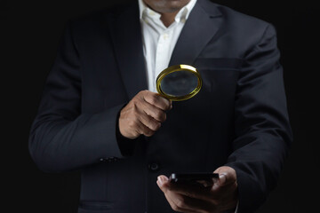 business man holding magnifier and smart phone against dark background.