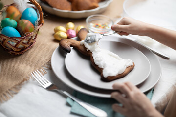 Childs hands decorating Easter bunny shaped cookie with icing. Multi colored Easter eggs in wicker basket and chocolate eggs on festive decorated table - obrazy, fototapety, plakaty