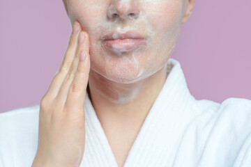 Young woman applying cleansing foam on face close-up on lilac background.