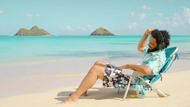 African American Young guy with the headphones and the laptop sitting on a beach chair at the island and working remotely, happy excited talking on the zoom. Remote work, outside office in Paradise