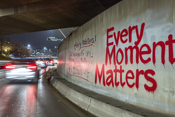 Graffiti on a city underpass wall reading 'Every Moment Matters' with blurred traffic in motion