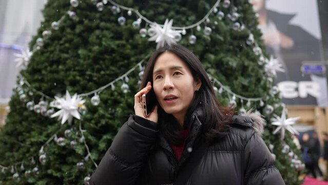 韓国のソウルの明洞の寒い冬にクリスマスツリーの前でスマートフォンで通話する30代の韓国人男性のスローモーション映像 Slow-motion video of a Korean man in his 30s talking on his smartphone in front of a Christmas tree in the cold winter in Myeong-dong, Seoul, 