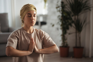 Young woman with cancer taking yoga and meditating in her apartment. Strong female patient calming...