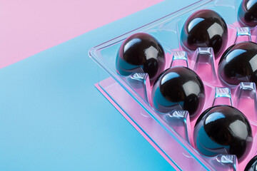 Black shiny modern plastic Easter eggs in a translucent  egg carton tray in a maximalist, surrealistic, and retro futuristic art style, asymmetrical, vaporwave composition in pink, blue background