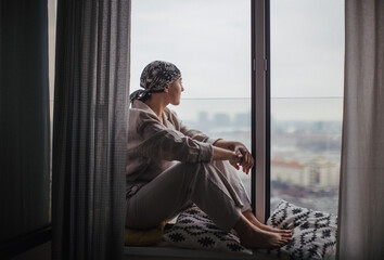 Young unhappy woman with cancer sitting in a window and looking at view.