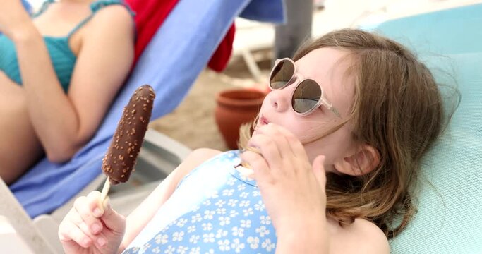 Portrait of cute little girl holding chocolate ice cream. Child in sunglasses enjoying sweet treat lying on sun lounger outdoors