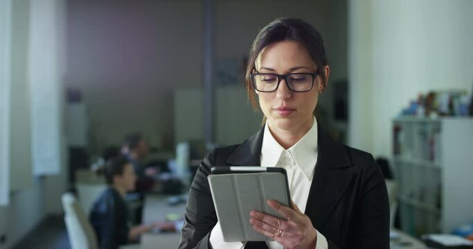 Portrait of Successful Businesswoman in Formal Clothes Using Tablet Computer, Standing in Modern Diverse Office Working on Financial, Business and Marketing Projects. She Looks at Camera and Smiles