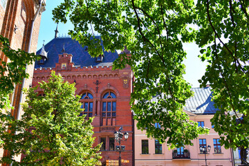 Old buildings made of red brick, decorative elements on the facade, high arched windows and blue roof. Green branches of the trees around. Torun, Poland, August 2023 