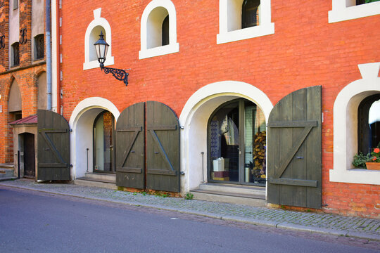 Facade of the historic building with small semicircular windows, antique doors with wooden sliding shutters, lantern, red brick wall. Ancient architecture, old street. Torun, Poland, August 2023 