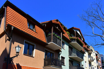 Facade of ancient building with wooden balconies and flowers, low windows, red tiled roof and street lamp. Blue sky and the branches of a tree without leaves. Torun, Poland, August 2023 