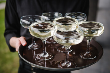 Tray of glasses of summer sparkling wine for guests at wedding reception. The waiter serving champagne on tray. Waiter holding glasses in the backyard. Man brings full glasses of champagne on a tray