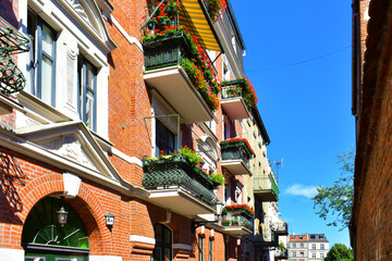 Facade of ancient red brick building, decorative elements, arch, balconies with wrought iron elements and flowers, a street lamp. Blue sky. Torun, Poland, August 2023 