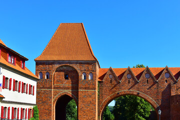 Medieval brick wall with tower, red tiled roof and arched passage. Ancient architecture, old castle. Summer sunny day with blue sky. Poland, Torun, August 2023.