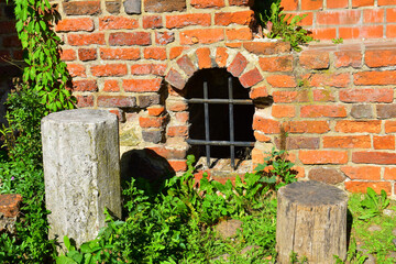Old red brick wall and window with a grid. An abandoned old building. Castle Ruins. Grass and plants around. Torun, Poland, August 2023