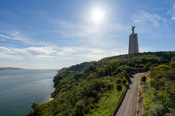 statue of Christ in Almada/Lisbon, with the morning sun falling on the tarred road and the Tagus River - obrazy, fototapety, plakaty
