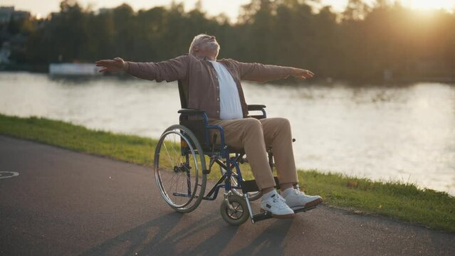 Senior man in wheelchair enjoys freedom. Male with special need spreading arms on sides smiling breathing fresh air walking in the city park. Living with physical disability in old age concept.