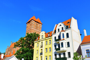 Ancient buildings with colorful walls, tower and red roof, made of red brick. Ancient architecture, old street. Green trees and blue sky. Torun, Poland, August 2023 