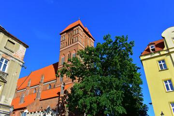 The tower ancient building with long windows, decorative elements and red roof, made of red brick. Ancient architecture. Green trees and blue sky. Torun, Poland, August 2023 