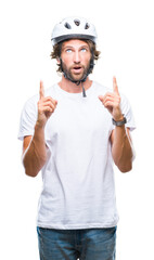 Handsome hispanic cyclist man wearing safety helmet over isolated background amazed and surprised looking up and pointing with fingers and raised arms.