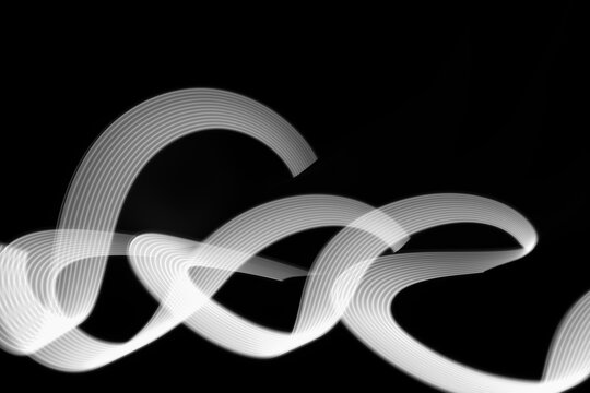 White neon curved wave of light as curls or swirl with smooth stripes on black background, pattern. Abstract background with motion light effect, light painting in New Year style.