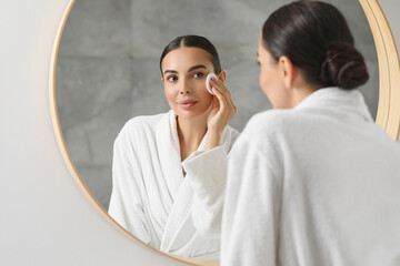 Beautiful woman removing makeup with cotton pad near mirror indoors