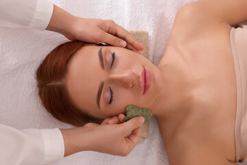Obraz na płótnie Canvas Young woman receiving facial massage with jade gua sha tool in beauty salon, above view