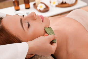 Young woman receiving facial massage with jade gua sha tool in beauty salon, closeup