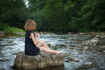 A young girl sits on a stone on a mountain river
