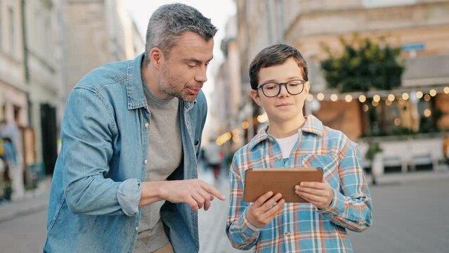 Frontal view of Caucasian father teaching his son how to use digital device in middle of street. Father pointing at something in distance and child taking photo with tablet while smiling.